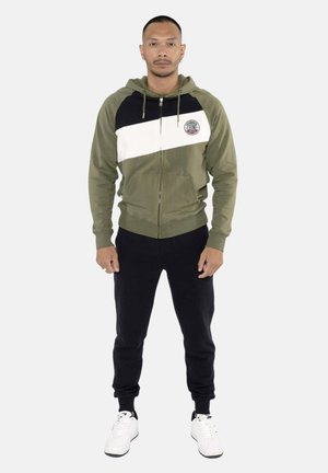 Homme debout face à l'avant portant un sweat à capuche zippé vert olive, noir et blanc, pantalon noir et baskets blanches sur un fond uni.