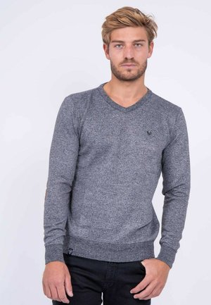 Homme aux cheveux blonds et à la barbe, portant un pull gris ajusté à col en V et un pantalon noir, se tenant devant un fond blanc uni.