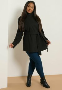 Mujer con cabello largo y oscuro, vistiendo un abrigo negro con cinturón, jeans azul oscuro y botas negras de tobillo, de pie contra una pared blanca.