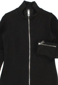 Giacca nera con zip e colletto alto, con una chiusura lampo argentata lucida e una piccola tasca con zip sulla manica sinistra. Texture liscia.