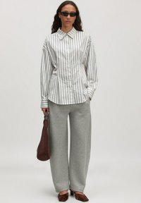 Wit en zwart gestreepte button-up shirt met een kraag, gecombineerd met grijze wijde sweatpants. Accessoires zijn een bruine tas en zwarte zonnebril.