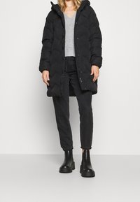 Manteau matelassé noir avec capuche, présentant un design matelassé. Porté sur un pull gris et un jean noir, accompagné de bottes noires épaisses.