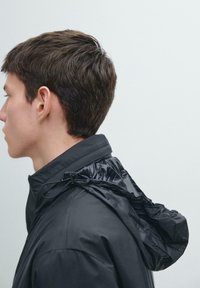 Schwarze, leichte Jacke mit Kapuze und Kordelzug, glatter Textur und strukturiertem Kragen. Seitenansicht des Modells gezeigt.