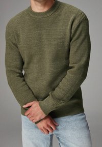 Man in een olijfgroene geribde trui en lichtblauwe jeans, met de handen gekruist op de taille tegen een grijze achtergrond.