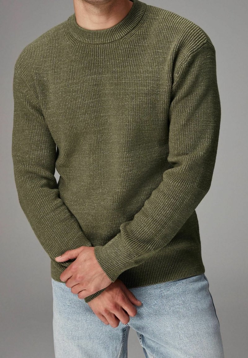 Man in een olijfgroene geribde trui en lichtblauwe jeans, met de handen gekruist op de taille tegen een grijze achtergrond.