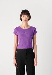 Purple ribbed short-sleeve top with a round neckline and a black logo patch on the chest, paired with black high-waisted trousers.