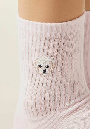 Blassrosa gerippte Socke mit einem kleinen weißen, gestickten Hundegesicht am Knöchel, getragen an einem nackten Bein vor neutralem Hintergrund.