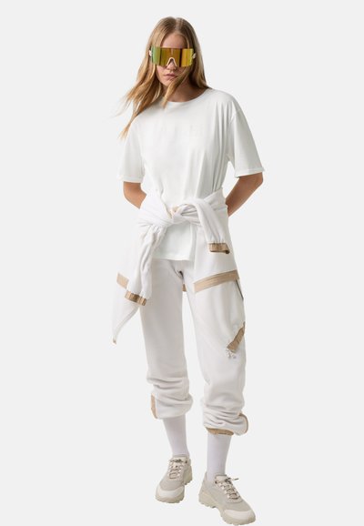 Žena v bílé oversized tričku, bílých joggerách s béžovými akcenty, bílých teniskách a velkých reflexních slunečních brýlích, s rukama za zády.