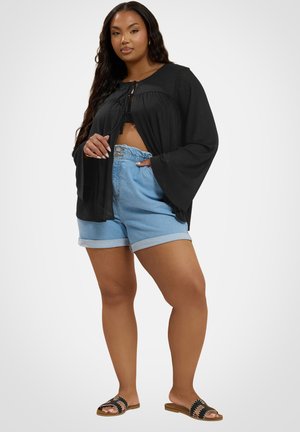 Krągła kobieta stoi, ubrana w luźną czarną bluzkę z długim rękawem, jasnoniebieskie podwinięte szorty jeansowe oraz czarne wsuwane sandały.