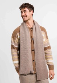 Homme souriant portant un cardigan rayé marron et une écharpe beige, se tenant contre un fond clair uni.