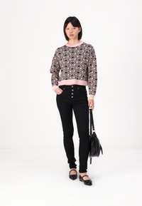 Gepflegte rosa Sweatshirt mit schwarzem Herzmuster, kombiniert mit hochgeschnittenen schwarzen Jeans und schwarzen Ballerinas. Das Model hält eine schwarze Handtasche.