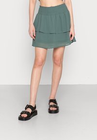 Falda en capas de color verde azulado con cintura fruncida y material de gasa translúcida. Combinada con sandalias negras con hebillas y suela plataforma.