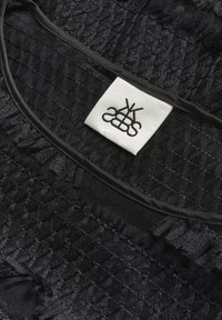 Melnā tekstūra audums ar sakratītu šuvju, un tuvāk melnajam apmalei ir šūta balta kvadrātveida etiķete ar stilizētu melnu logo.