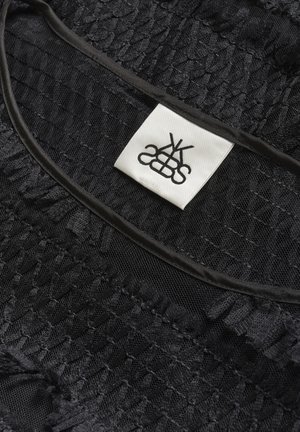 Tissu noir texturé avec des coutures froncées et une étiquette carrée blanche comportant un logo noir stylisé cousue près d'un bord noir.