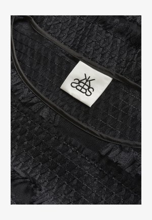 Tissu noir texturé avec des coutures froncées et une étiquette carrée blanche comportant un logo noir stylisé cousue près d'un bord noir.