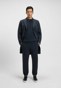 Navy checkered long coat over a dark sweater and matching tapered pants. Black sneakers with white soles. Smooth, tailored fabric.