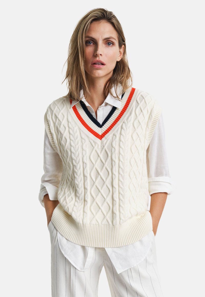 Gilet lavorato a maglia color crema con scollo a V e fondo a costine, con accenti a strisce arancioni e navy. Indossato sopra una camicia bianca e pantaloni a righe.