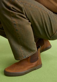 Brown leather ankle boots with a smooth finish, elastic side panels, and rubber soles, paired with dark green textured pants.