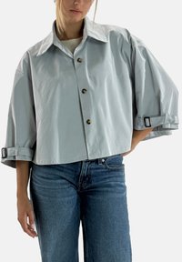 Lysegrå cropped jakke med stor krave, knaplukning foran og opkørte ærmer, der holdes på plads med stropper. Glat stof med en afslappet pasform.