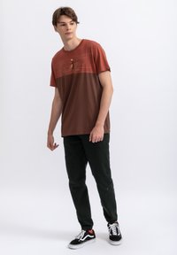 Bruine en roestkleurige t-shirt met een grafisch ontwerp, gecombineerd met donkergroene broek. Model draagt zwart-witte sneakers. Eenvoudige, casual outfit.