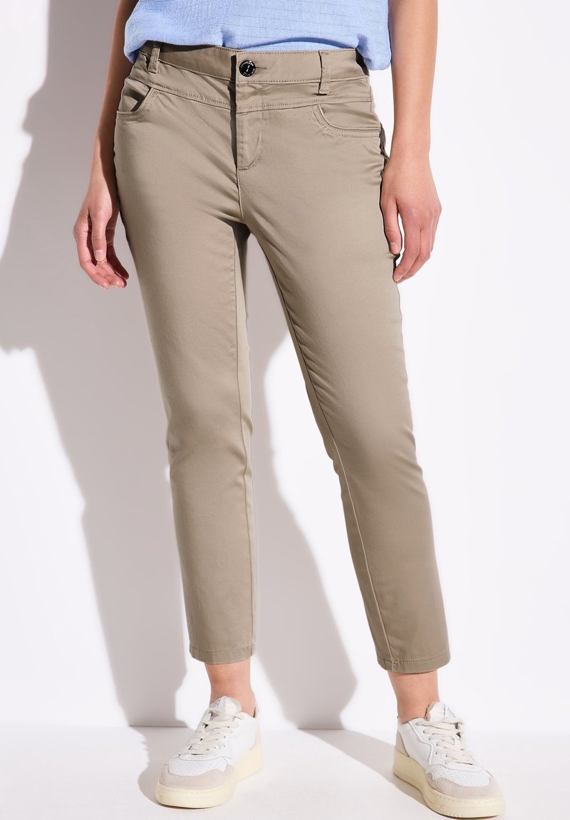 Femme portant un pantalon beige ajusté et court, un haut bleu clair et des baskets blanches, posant devant un fond blanc.