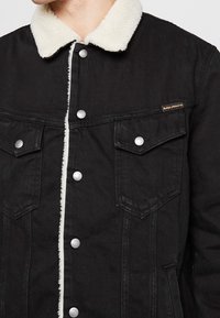 Svart denimjacka med en krämfärgad shearlingkrage, silverfärgade tryckknappar och två bröstfickor med spetsiga lock.