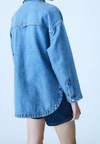 Světlomodrá džínová košile s dlouhými rukávy, volným střihem, zaobleným lemem a knoflíkovými manžetami, ve spojení s tmavými džínovými šortkami.