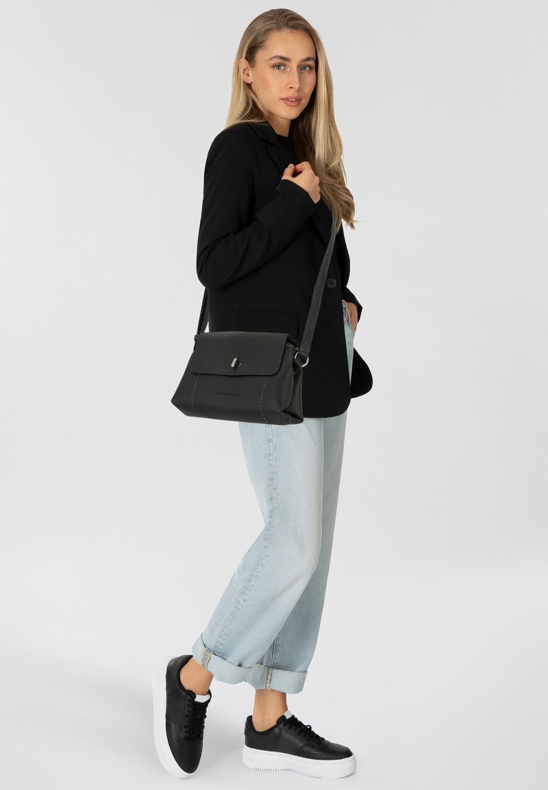 Borsa a tracolla nera realizzata in materiale testurizzato, con una patta dotata di una chiusura in metallo, indossata da una persona in giacca nera e jeans di denim chiaro.