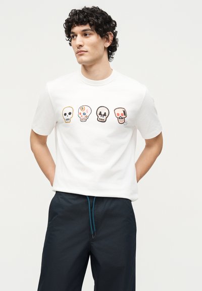 Νέος άνδρας με σγουρά μαλλιά που φοράει λευκό μπλουζάκι με τέσσερα πολύχρωμα, στυλιζαρισμένα κρανία στο στήθος, συνδυασμένο με σκοτεινό παντελόνι.