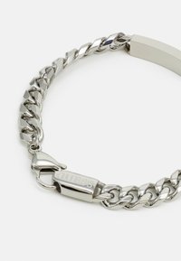 Μεταλλικό βραχιόλι με χοντρή αλυσίδα σχεδίασης, με λειασμένους ασημένιους κρίκους, κούμπωμα που φέρει την ένδειξη "GUESS" και ομαλό, επίπεδο λουρί.