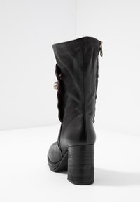 Svart läderstövel till mitten av vaden med staplad blockklack, sidodragkedja och dekorativt spänne, fotograferad mot en vit bakgrund.