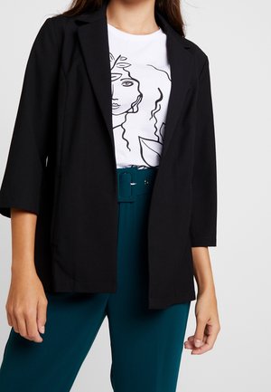 Frau trägt schwarzen offenen Blazer, weißes T-Shirt mit schwarzem Linienkunst-Gesichtsprint und petrolfarbene hoch taillierte Hose mit Gürtel vor grauem Hintergrund.