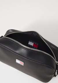 Černá kožená toaletní taška se stříbrným zipem, uvnitř s označením značky "TOMMY JEANS" a malým logem na vnějším povrchu.
