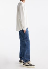 Vit långärmad tröja med rund hals, kombinerad med blå cargobyxor med sidfickor. Modellen bär svarta och vita sneakers.