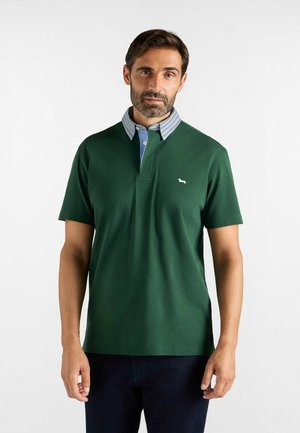 Uomo con capelli castani corti e barba, indossa una polo verde a maniche corte con colletto a righe e pantaloni scuri, in piedi su sfondo bianco.