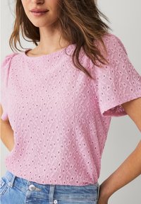Lyserød kortærmet top lavet af struktureret stof med blomsterbroderi. Har en rund halsudskæring og bløde puffede ærmer. Bæres med blå jeans.