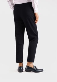 Pantaloni neri su misura con taglio dritto, orlo con risvolto e tasche laterali, abbinati a mocassini scuri lucidi su uno sfondo chiaro.