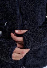 Giacca in pile blu navy con superficie testurizzata, zip intera e polsini elastici. Presenta due tasche con zip e una sensazione morbida e accogliente.