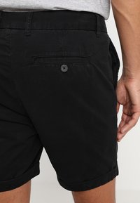 Schwarze Baumwollshorts mit geradem Schnitt, ausgestattet mit einer geknöpften Gesäßtasche und umgeschlagenen Säumen. Glatte Textur und minimale Designdetails.