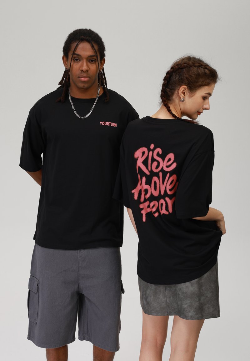 Černé oversized trička mají na přední straně logo "YOURTURN" v růžové barvě a na zadní straně nápis "Rise Above Fear" ve stylu štětce.