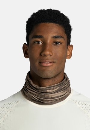 Giovane uomo con capelli corti e ricci che indossa una sciarpa marrone fantasia e una camicia bianca traforata, che guarda avanti con un'espressione neutra.