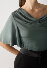 Top cu guler tip cowl într-un material verde stins; mâneci scurte și vaporoase; textură ușor transparentă; asortat cu pantaloni negri cu talie înaltă.