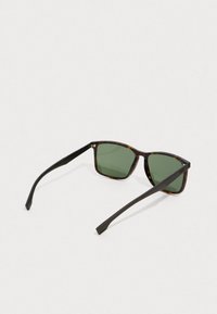 Kilpikonnakuosiset aurinkolasit, joissa on tummanvihreät suorakulmaiset linssit ja mustat suorat sivukahvat, lepäävät auki valkoisella pinnalla.