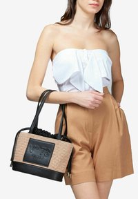 Beige gevlochten boodschappentas met zwarte leren accenten en logo. Voorzien van twee handvatten en een trekkoordsluiting. Draagt met een witte top en shorts.