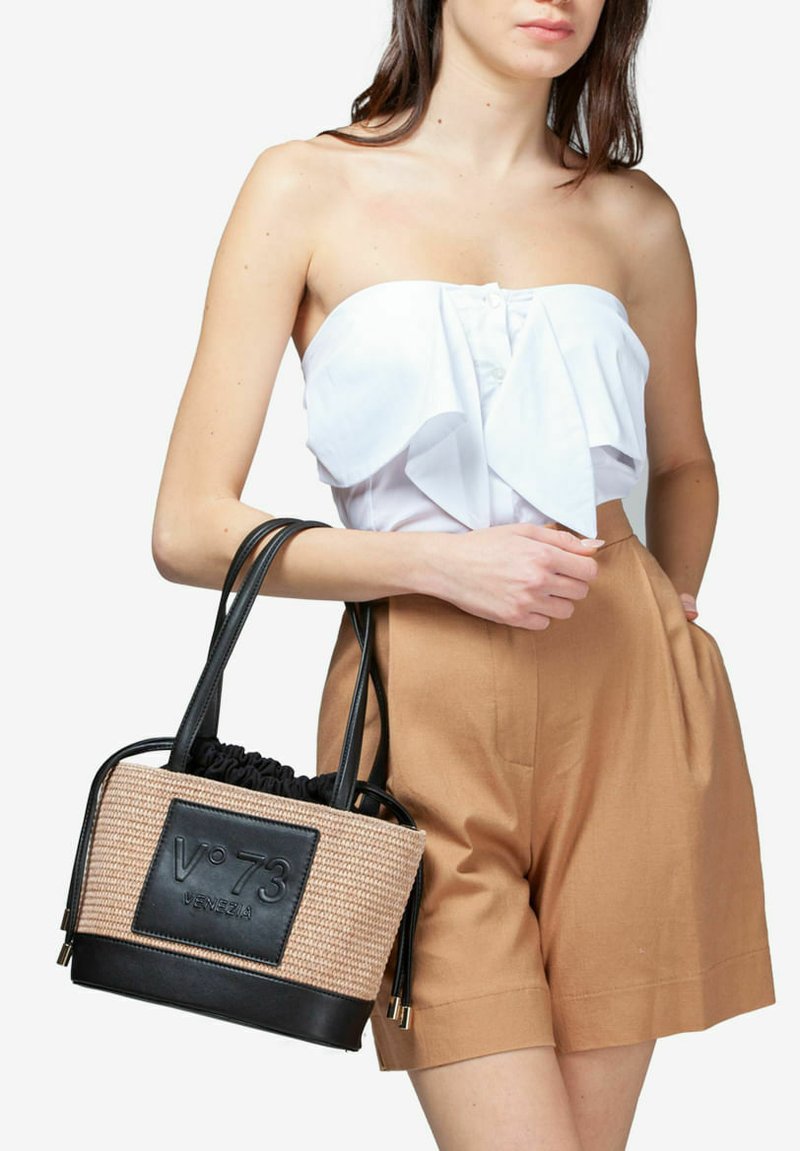 Beige gevlochten boodschappentas met zwarte leren accenten en logo. Voorzien van twee handvatten en een trekkoordsluiting. Draagt met een witte top en shorts.