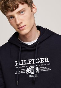 Sudadera color azul marino hecha de algodón, con un estampado de logo blanco y cordones con puntas de metal. Textura suave, diseño casual.