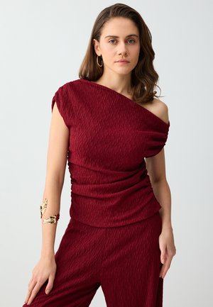 Kobieta o brązowych włosach sięgających ramion, ubrana w teksturowany bordowy top na jedno ramię oraz dopasowane spodnie, uzupełniona złotymi kolczykami kołami oraz bransoletką na rękę.
