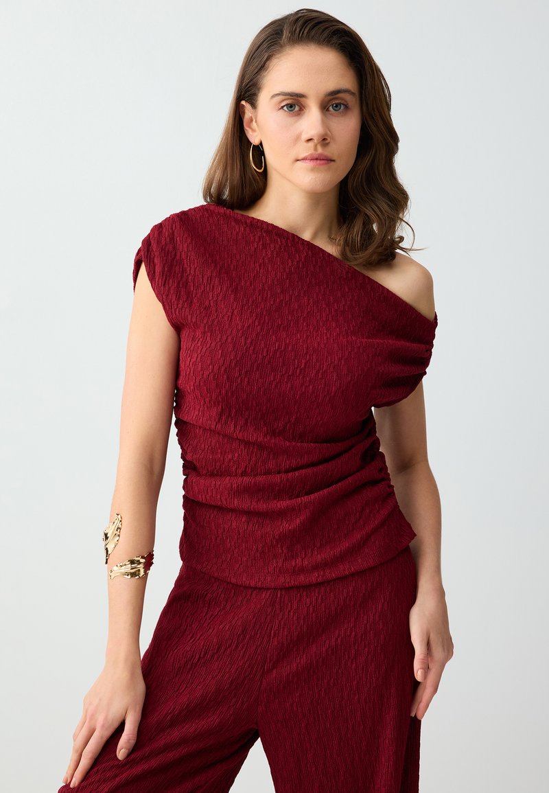 Femme aux cheveux bruns mi-longs portant un top texturé bordeaux à une épaule et un pantalon assorti, accessoirisée avec des boucles d'oreilles créoles dorées et un bracelet de bras.