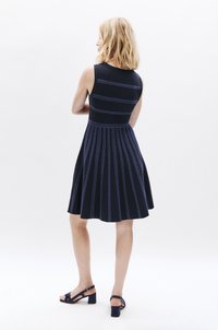 Marineblaues Kleid mit ausgestelltem Rock, versehen mit vertikalen blauen Streifen und einem taillierten Oberteil. Ein Paar marineblaue Absatzsandalen rundet den Look ab.
