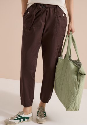 Personne portant un pantalon marron foncé, des baskets blanches avec des rayures vertes, tenant un sac cabas matelassé vert clair par les poignées.